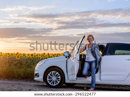 Smiling Young Female Driver in Trendy Outfit, Getting Out from her White Car and Looking at the Camera Against Sunflower Field.