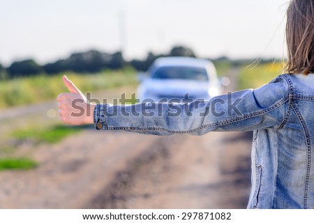 Young woman hitchhiking in the countryside flagging doan an approaching car on a farm road with her thumb