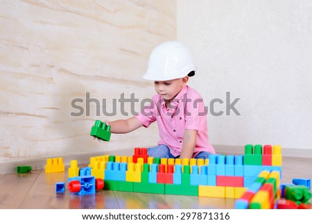 Young boy wearing hardhat playing indoors sitting on a wooden floor grinning as he holds up a large colorful plastic building block