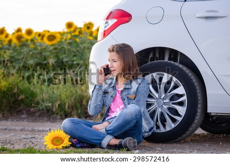 Young woman chatting on her mobile phone as she leans against the car tyre while parked in a rural road near a field of sunflowers