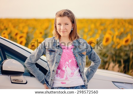 Young girl standing leaning on the bonnet of a car parked in the countryside in front of a field of sunflowers