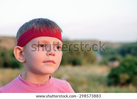 Close up of the face of a handsome young boy wearing a red headband staring into the distance into the setting sun with a dreamy expression