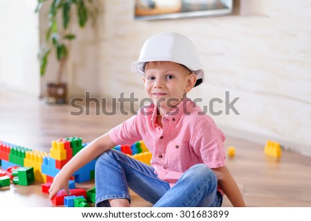 Happy little boy sitting on the floor and wearing plastic white hard hat, blue jeans and pink shirt while playing with toy construction blocks, at home