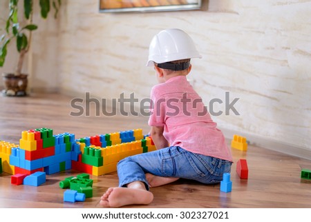 Young boy wearing hardhat playing indoors sitting on a wooden floor grinning as he holds up a large colorful plastic building block