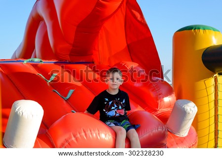 Blonde boy (7-9 years) wearing t-shirt and multi-colored shorts sitting in red bouncy castle, blue sky in background