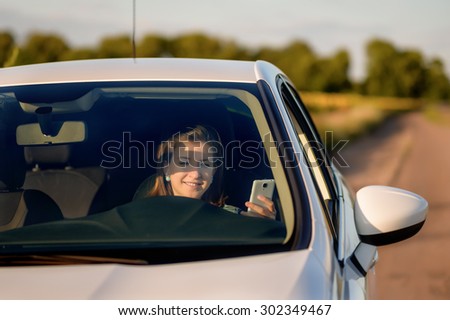 Woman driver smiling as she reads an sms or text message on her mobile phone distracting her attention from the road, view through the windshield