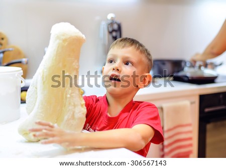 Young boy playing with dough while baking covering his arms in the uncooked mixture with a funny expression on his face as he stands at the kitchen counter