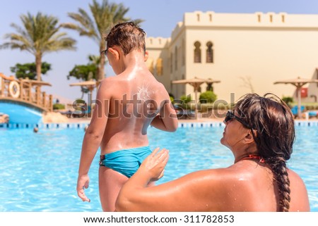 Young mother rubbing sunscreen onto her son as he stands in the summer sunshine over looking a sparkling swimming pool at a tropical resort