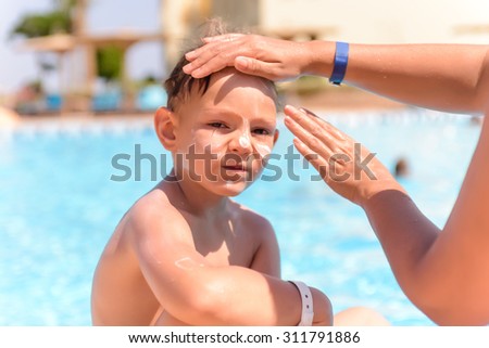 Woman applying sunscreen to the face of her young son as he sits waiting to go swimming alongside a swimming pool, close up of him looking at the camera