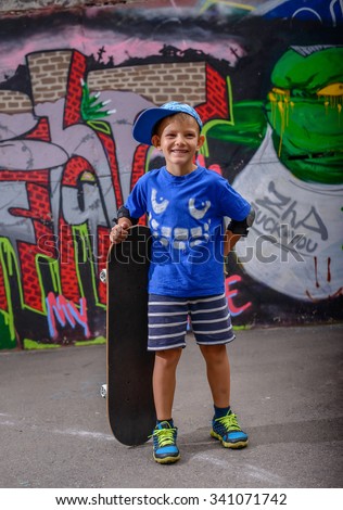 Happy young boy posing with his skateboard in a trendy blue outfit in front of a colorful wall painted with graffiti laughing at the camera