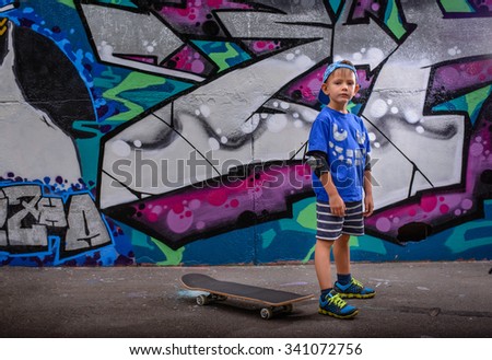 Little boy standing in front of vibrant graffiti in a trendy blue outfit looking at the camera with his skateboard on the ground behind him