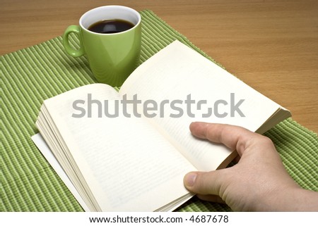 Hand holding a book on a table with a green coffee cup. The book has blank pages.