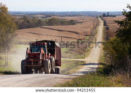 Rural landscape with a tractor pulling a trailer on foreground and hilly corn fields with a gravel road on background.