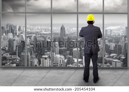 rear view engineer standing cross one\'s arm and concrete interior of modern area at the top floor high building with urban scene balcony over looking city dusky before rain falling