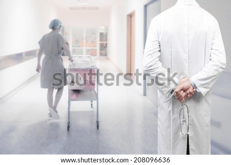 rear view image of doctors with stethoscope in a hospital pose arms crossed behind back looking at female nurse pushing stretcher gurney bed in labour room of hospital corridor with baby