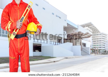 technician in uniform holding hard hat and yellow construction spirit level at the blurred modern building
