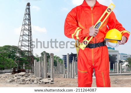 technician in uniform holding hard hat and yellow construction spirit level at the blurred construction site