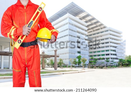 technician in uniform holding hard hat and yellow construction spirit level at the front of blurred modern building