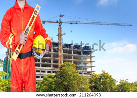 technician in uniform holding hard hat and yellow construction spirit level at the blurred construction background in blue sky