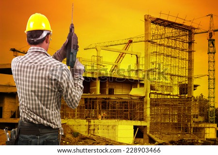 technician in protective safety equipment goggles hard hat with drill and tool belt  against building construction crane with beautiful sunset