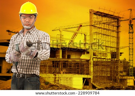 Portrait of technician in protective safety equipment goggles hard hat with drill  against building construction crane with beautiful sunset