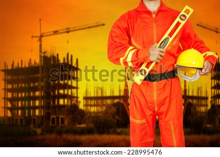 technician in uniform holding hard hat and yellow construction spirit level working at high building construction site against beautiful sunset
