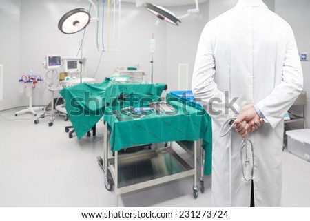 rear view image of doctors pose arms crossed behind back with stethoscope looking at operating room of a modern hospital