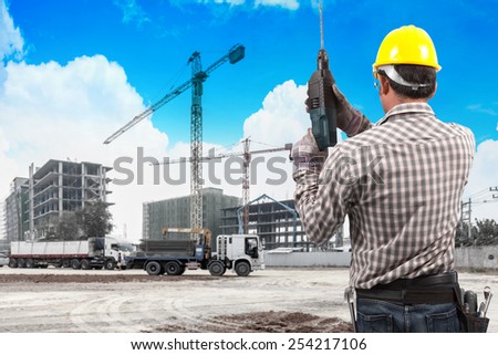 technician in protective safety equipment with drill and tool belt working in building construction site with tower crane and blue sky