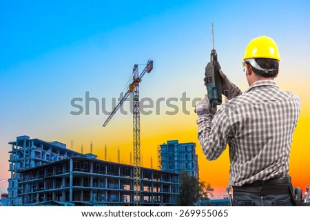 technician in protective safety equipment with drill and tool belt working at building construction site against beautiful sky sunset