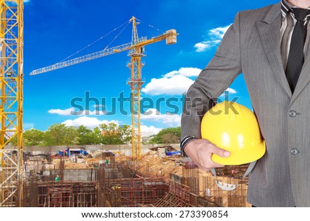 business man holding helmet for working at footing of building construction site with tower crane and blue sky