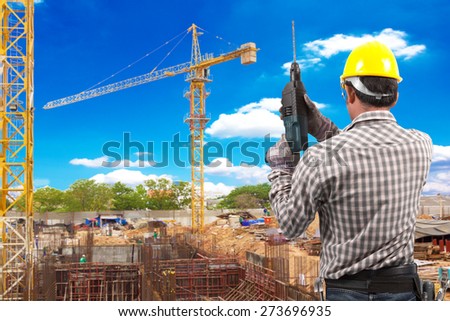 technician in protective safety equipment with drill and tool belt working at footing of building construction site with tower crane and blue sky