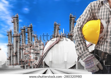 worker holding hard hat for working at pipe line connection to oil tanks in petrochemical oil refinery against blue sky