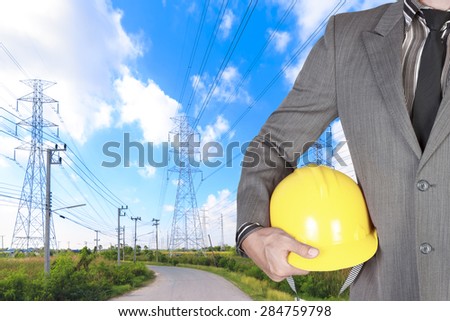 Engineer holding hard hat at high voltage power pylon against blue sky