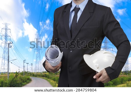 Engineer holding hard hat and blueprint for working at high voltage power pylon against blue sky