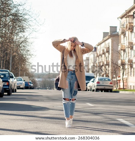 Adorable blonde girl walking alone on the road in old european city. Dressed on ripped jeans and coat