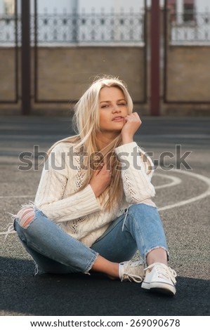 Young pretty fashionable blonde woman dressed in ripped jeans and white sweater sitting on the ground in summer and having fun