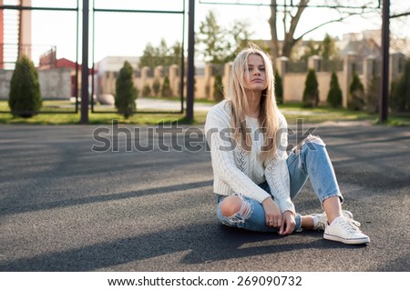 Young pretty fashionable blonde woman dressed in ripped jeans and white sweater sitting on the ground in summer and having fun
