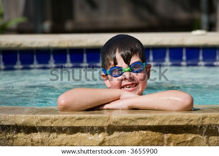 Young boy with arms crossed on side of swimming pool, wearing blue and green goggles