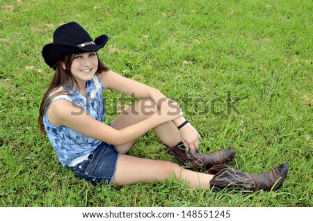 A young teenager with western hat and boots sitting in the grass smiling.