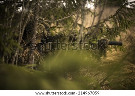 Camouflaged sniper keeps victim at gunpoint. Soldier dressed in ghillie camouflage on nature.
