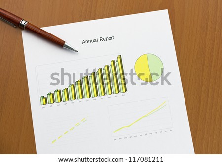 Annual report chart histogram, lines and area. Ink color print paper and a pen on wooden desk. Monthly stats.