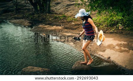 Barefoot happy indian woman in sunhat using stepping stones to cross the river.