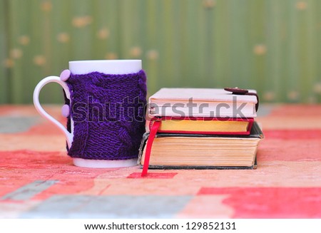 White mug with violet knitted sweater on it on the table with stack of books