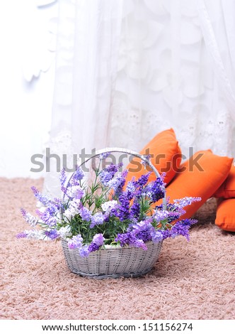 Basket with purple flowers on beige covered floor and orange pillows indoors