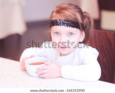 Cute baby girl holding cup of black tea in cafe