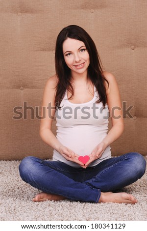 Smiling pregnant woman sitting on floor, holding little heart