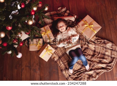 Kid girl 4-5 year old lying under christmas tree on wooden floor with christmas decorations, presents. Wearing trendy knitted sweater, jeans. Top view.