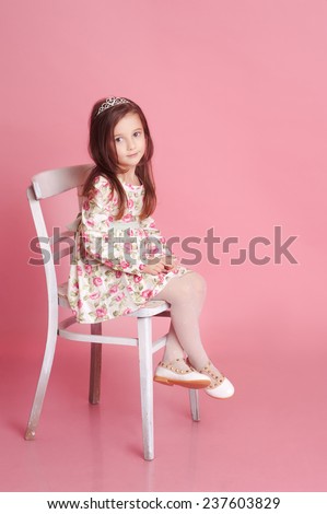 Smiling baby girl 4-5 year old posing in studio over pink. Sitting on chair. Wearing trendy floral dress.