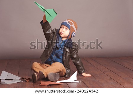 Little boy 4-5 year old playing with paper planes in room. Wearing stylish leather jacket and knitted pilot hat over gray.