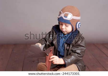 Cute baby boy wearing stylish leather jacket and aviator cap over gray. Playing with paper origami planes in room. Sitting on wooden floor. Childhood.